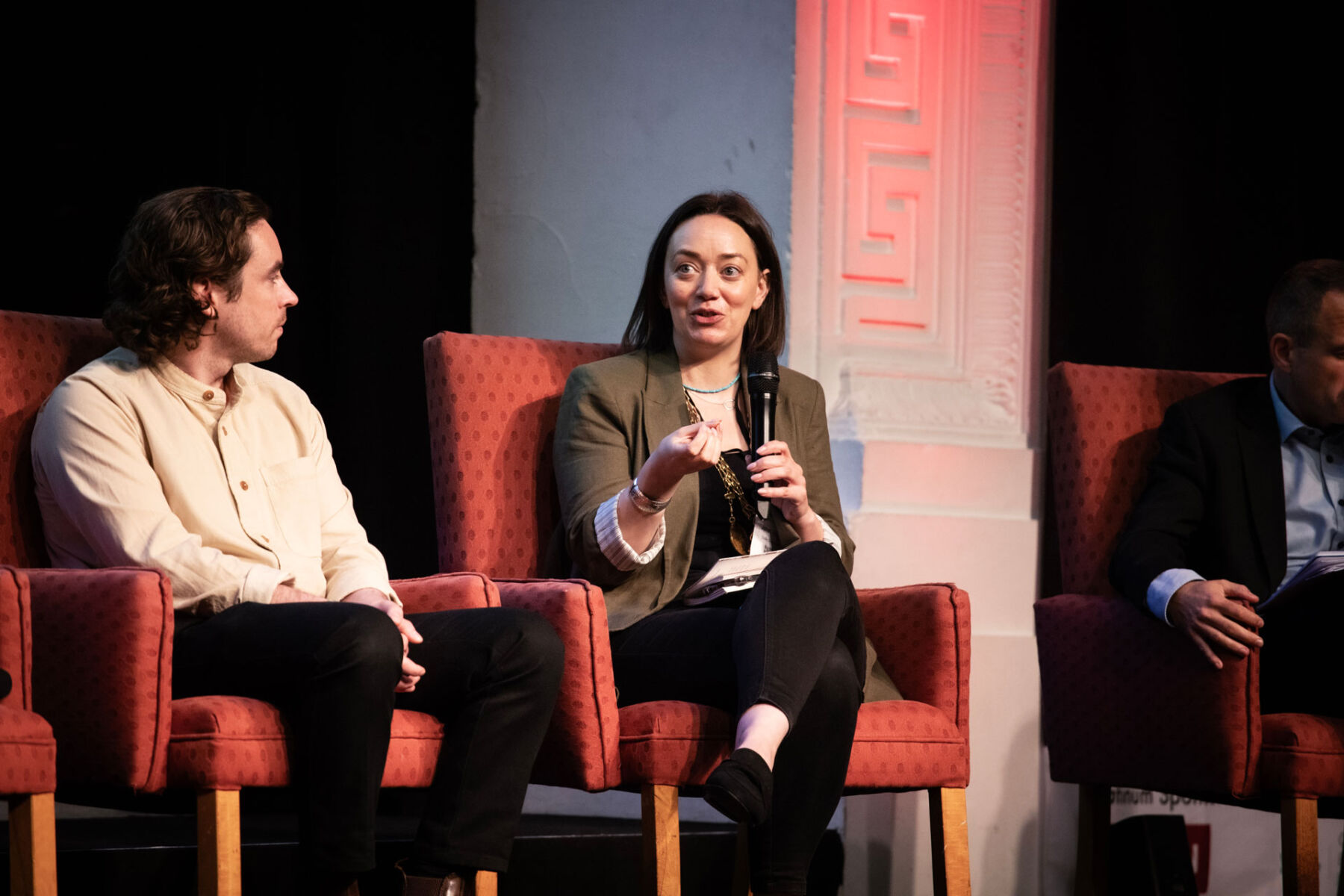 Panel discussion with three professionals, woman speaking with microphone, seated in red chairs.