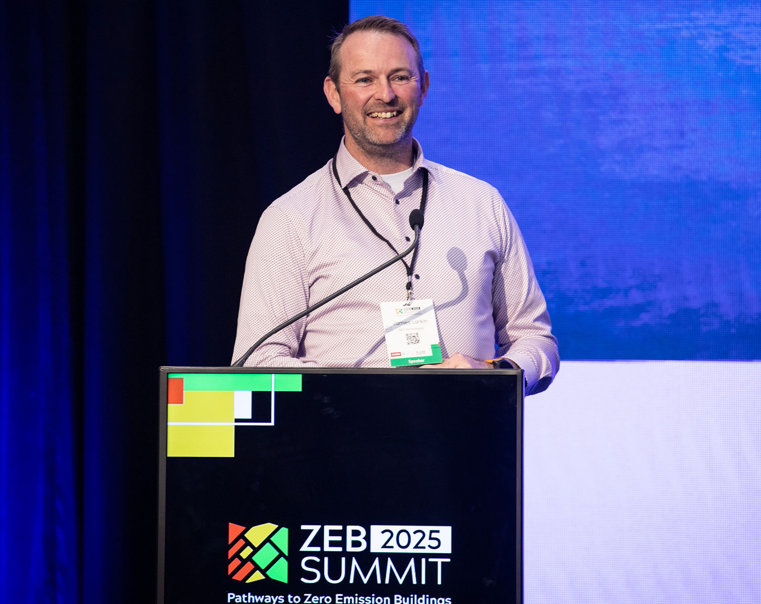 Speaker at ZEB 2025 Summit discusses zero emission buildings on stage with blue background.
