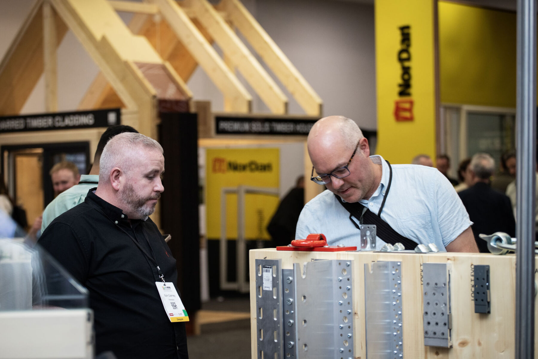 Two men discussing building materials at a construction trade show with wooden structures in the background.