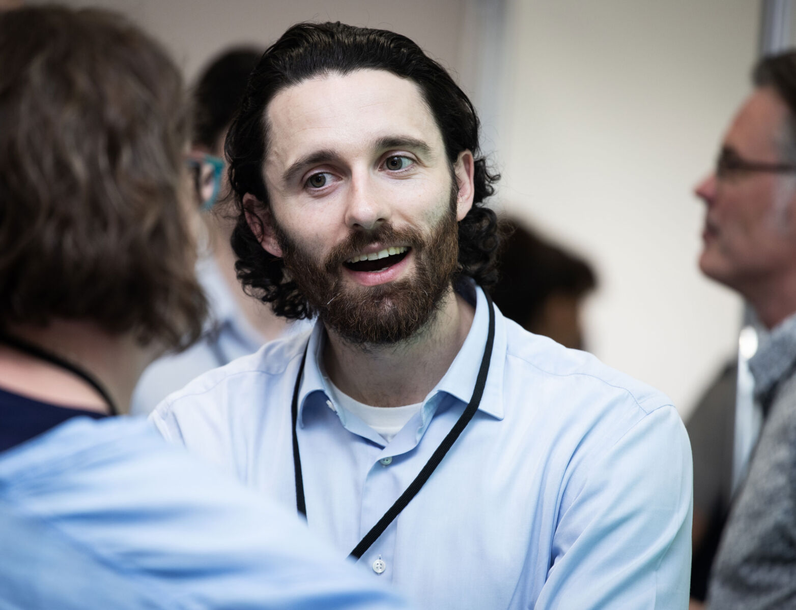 Person in conversation at networking event, wearing a light blue shirt and black lanyard, with others blurred in background.