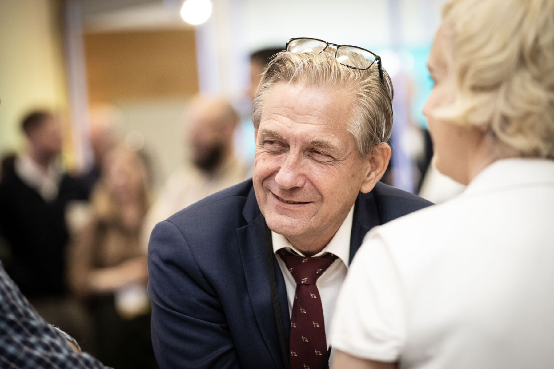 Smiling man in a suit at a social gathering, engaging in conversation with a group.