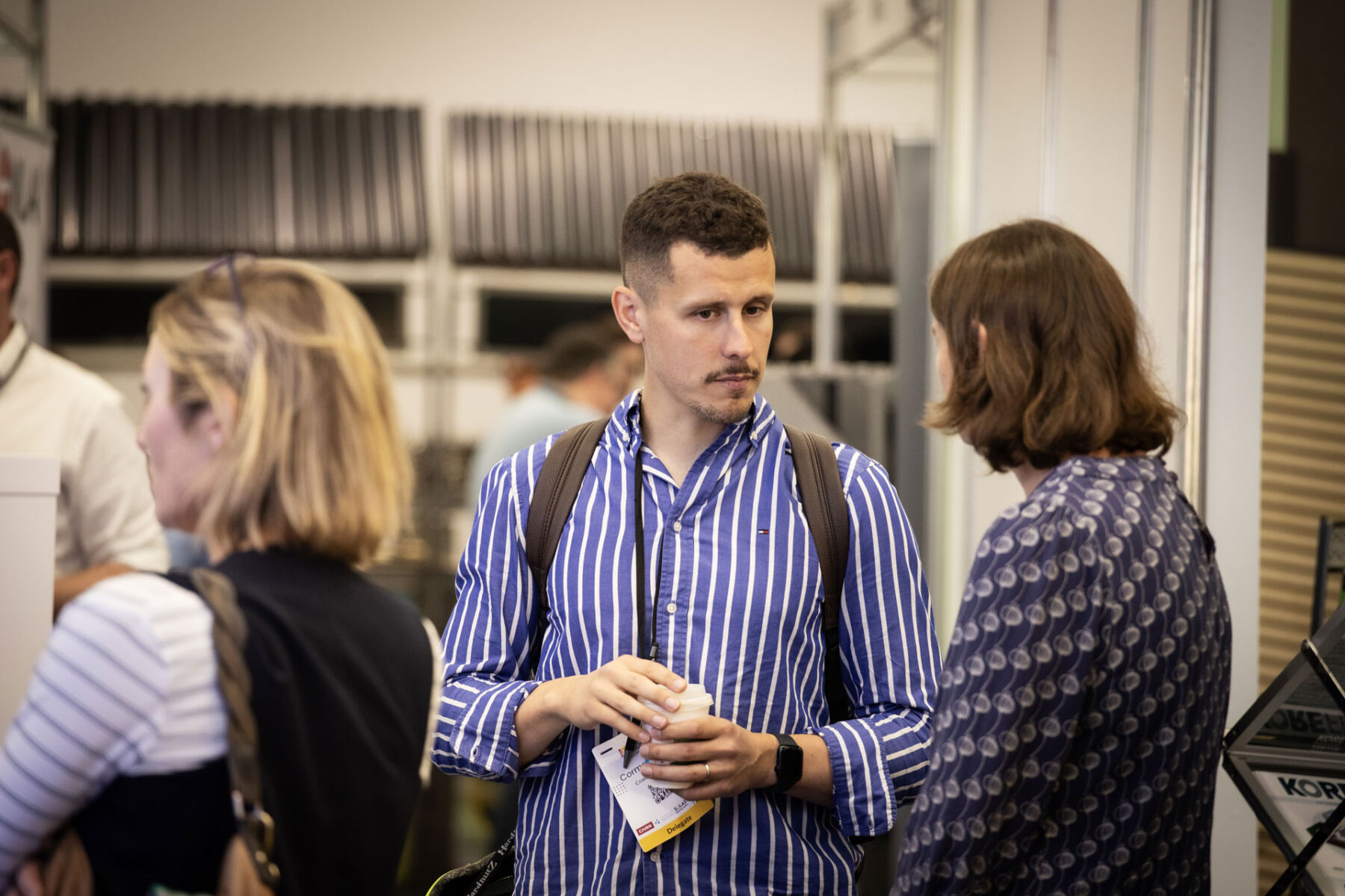 Man in blue striped shirt holding coffee cup, engaged in conversation at a conference.