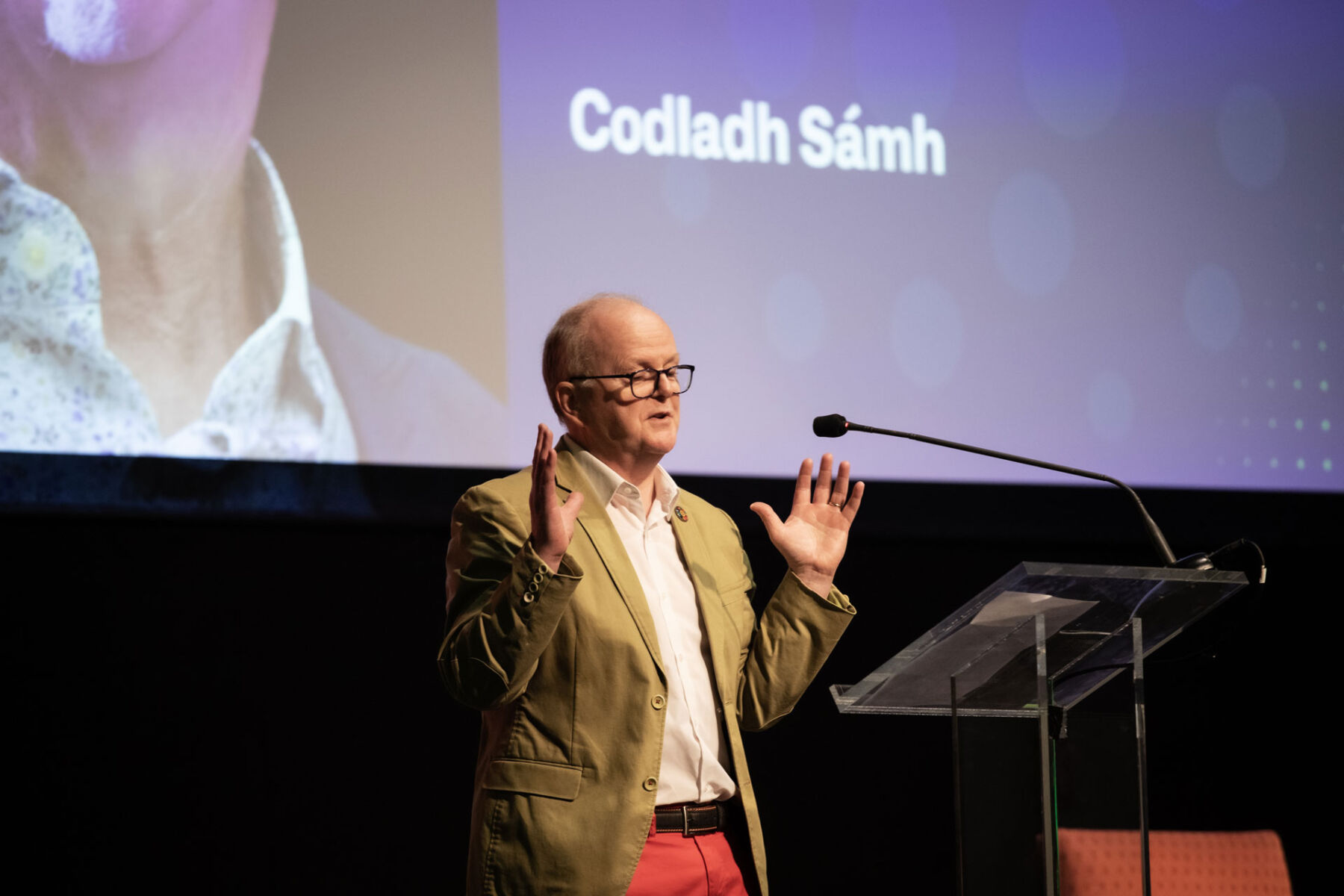 Speaker in a green jacket presenting on stage with Codladh Sámh in the background.