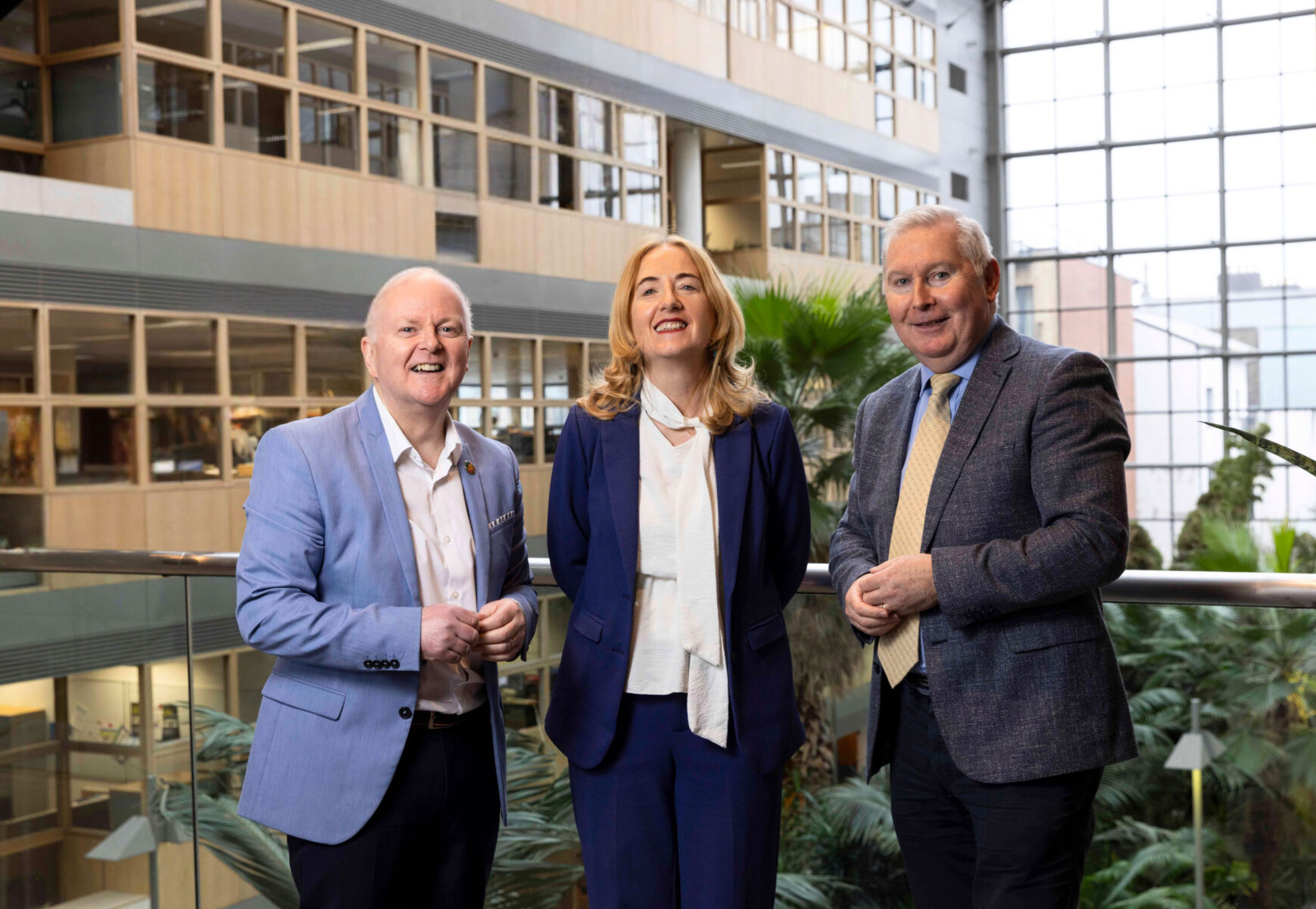 Three professionals standing in a modern atrium, smiling confidently near large windows and indoor plants.